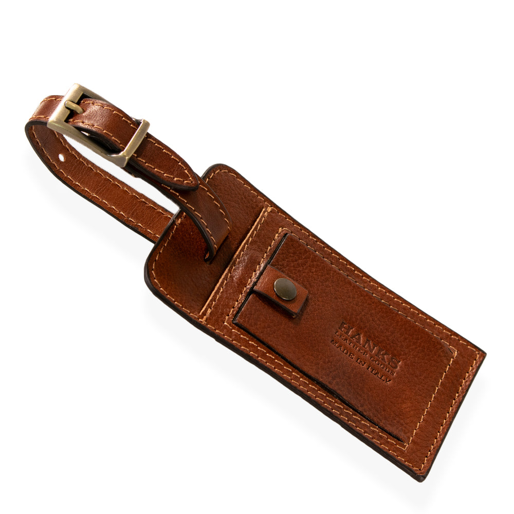 Introducing the Luxury Luggage Tag by Hanks Belts, meticulously crafted from premium Italian full-grain leather in a rich brown hue. This tag features a brass buckle and proudly displays embossed branding. It includes a front flap with a snap button closure that opens to reveal a slot for your personal information. The elegant stitching along the edges accentuates its classic and sophisticated appeal.