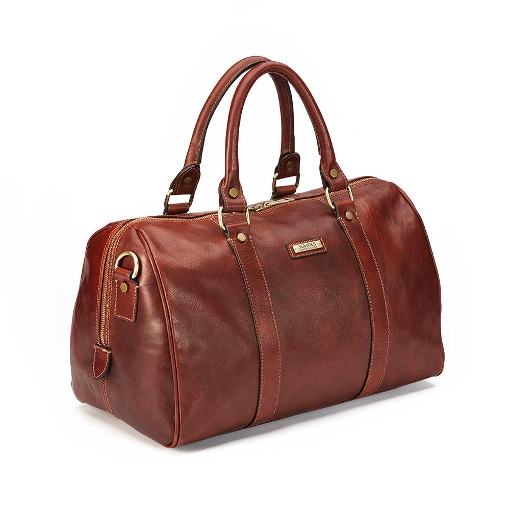 The Getaway Duffel With Luggage Sleeve from Hanks Belts is a stylish Italian leather travel bag in brown, featuring dual top handles and gold hardware such as a zipper and metal accents. This luxury duffel bag boasts a structured shape with a small logo plaque on the side and is carry-on-friendly for seamless journeys.