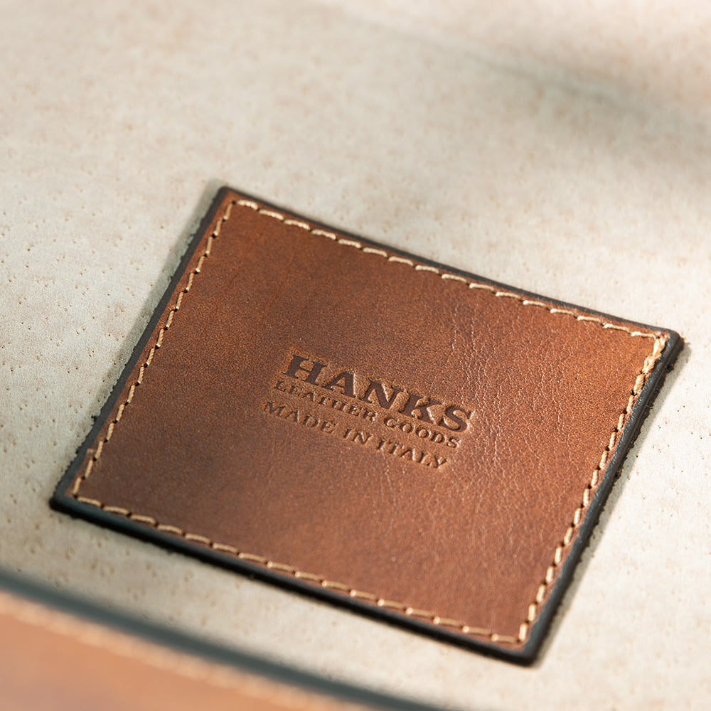 Close-up of a brown full-grain leather patch with stitching, engraved with the words &quot;HANKS BELTS MADE IN ITALY.&quot; The background is a soft, neutral fabric, perfectly showcasing the travel-ready elegance of the Italian Large Valet Tray.