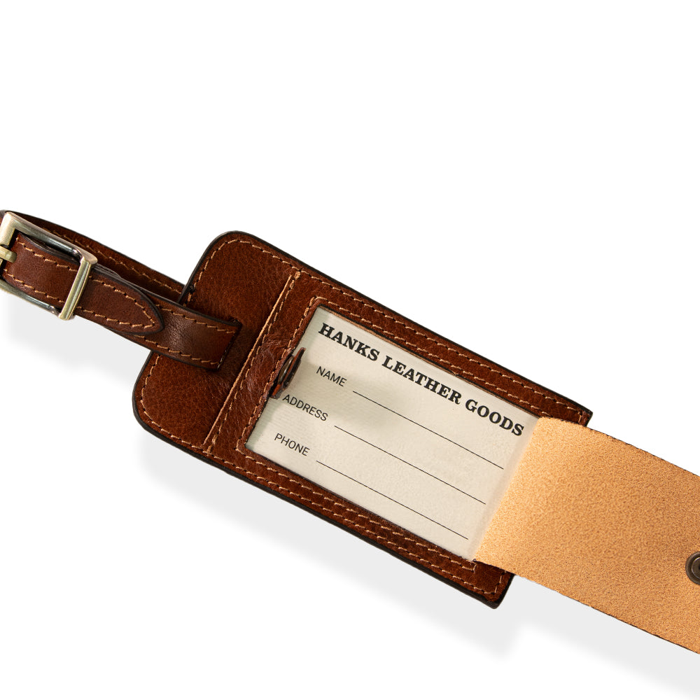 Introducing the Luxury Luggage Tag by Hanks Belts, meticulously crafted from premium Italian full-grain leather in a rich brown hue. This tag features a brass buckle and proudly displays embossed branding. It includes a front flap with a snap button closure that opens to reveal a slot for your personal information. The elegant stitching along the edges accentuates its classic and sophisticated appeal.