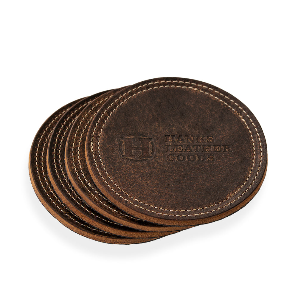 A stack of round, brown leather coasters with white stitching along the edges. The top coaster is embossed with &quot;Hanks Belts&quot; and a decorative logo. This set of four, known as the Premium Round Leather Coasters, is arranged in a slightly fanned-out manner.