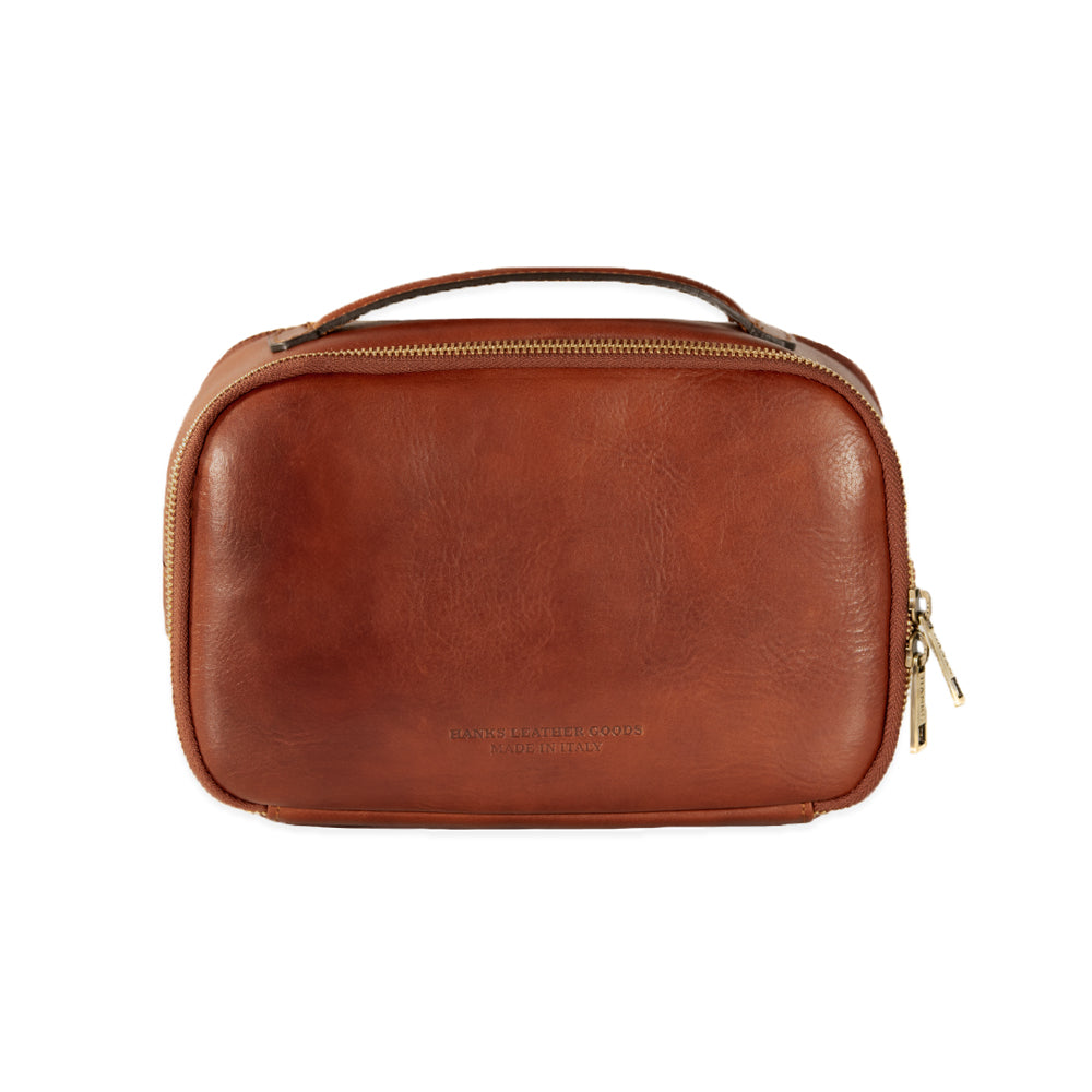 The Tidy Travel Case by Hanks Belts is a small, brown Italian leather toiletry bag featuring a zipper and a handle on top. The smooth-finish leather has subtle imprinted text on the front. With its gold-toned zippers and handle accents, this elegant travel case is perfect for organization.