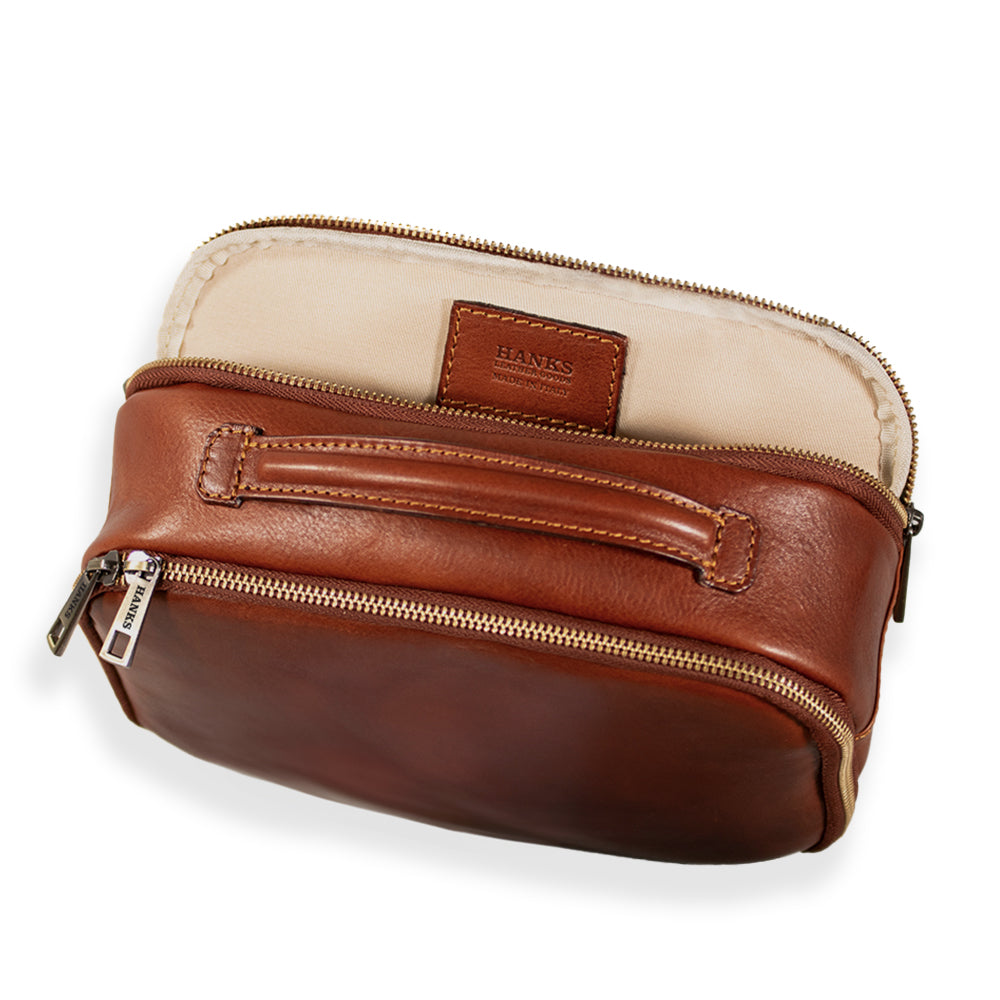 The Tidy Travel Case by Hanks Belts is a small, brown Italian leather toiletry bag featuring a zipper and a handle on top. The smooth-finish leather has subtle imprinted text on the front. With its gold-toned zippers and handle accents, this elegant travel case is perfect for organization.