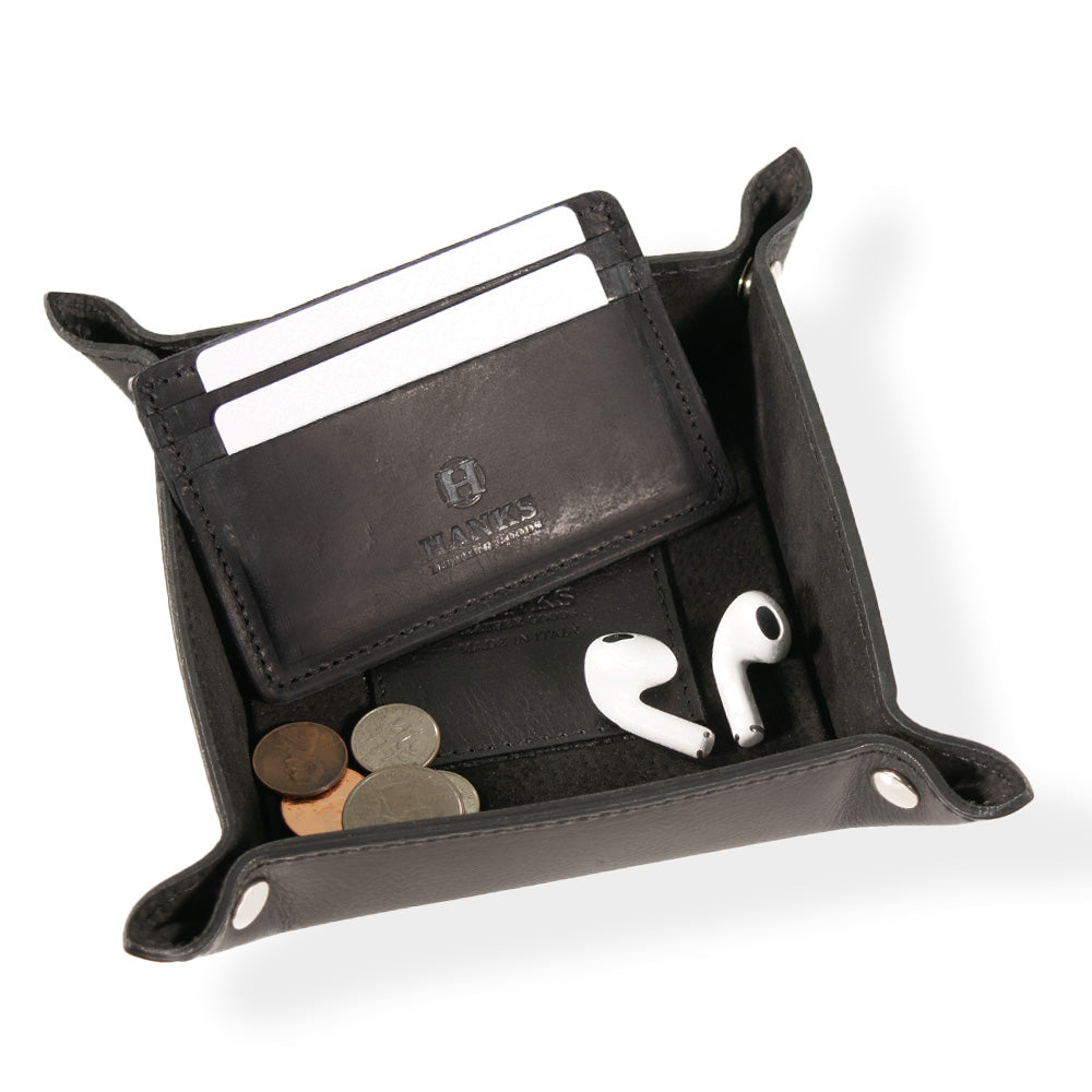 The Italian Compact Valet Tray by Hanks Belts is crafted from black Italian leather, featuring a compact rectangular shape with silver button snaps at each corner. The matching leather interior enhances its sleek look. Designed for travel and versatility, this tray is elegantly displayed on a plain white background.