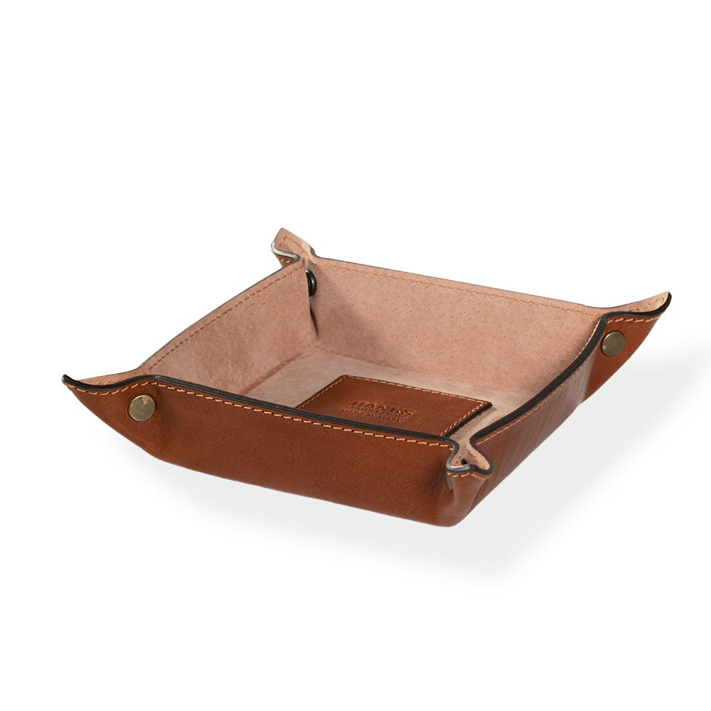 Introducing the Italian Compact Valet Tray by Hanks Belts: a sophisticated brown leather tray featuring visible stitching and brass fasteners at the corners. Its travel-friendly design includes a lighter leather lining inside, adorned with a small embossed logo on the base.