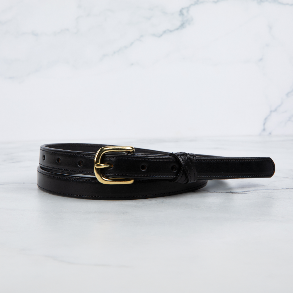 A Hanks Belts Dixie Belt - .75&quot; crafted from English bridle leather lies elegantly on marble. Its gold buckle enhances the rich black, with precisely crafted holes that showcase exceptional American craftsmanship against the white backdrop.