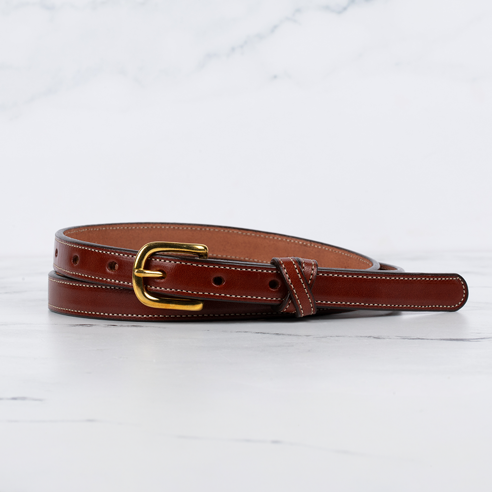 The Women&#39;s Dixie Belt by Hanks Belts, with a .75&quot; width, is crafted from premium English bridle leather featuring an oak color design, white stitching, and a gold buckle, elegantly displayed on a marble surface.