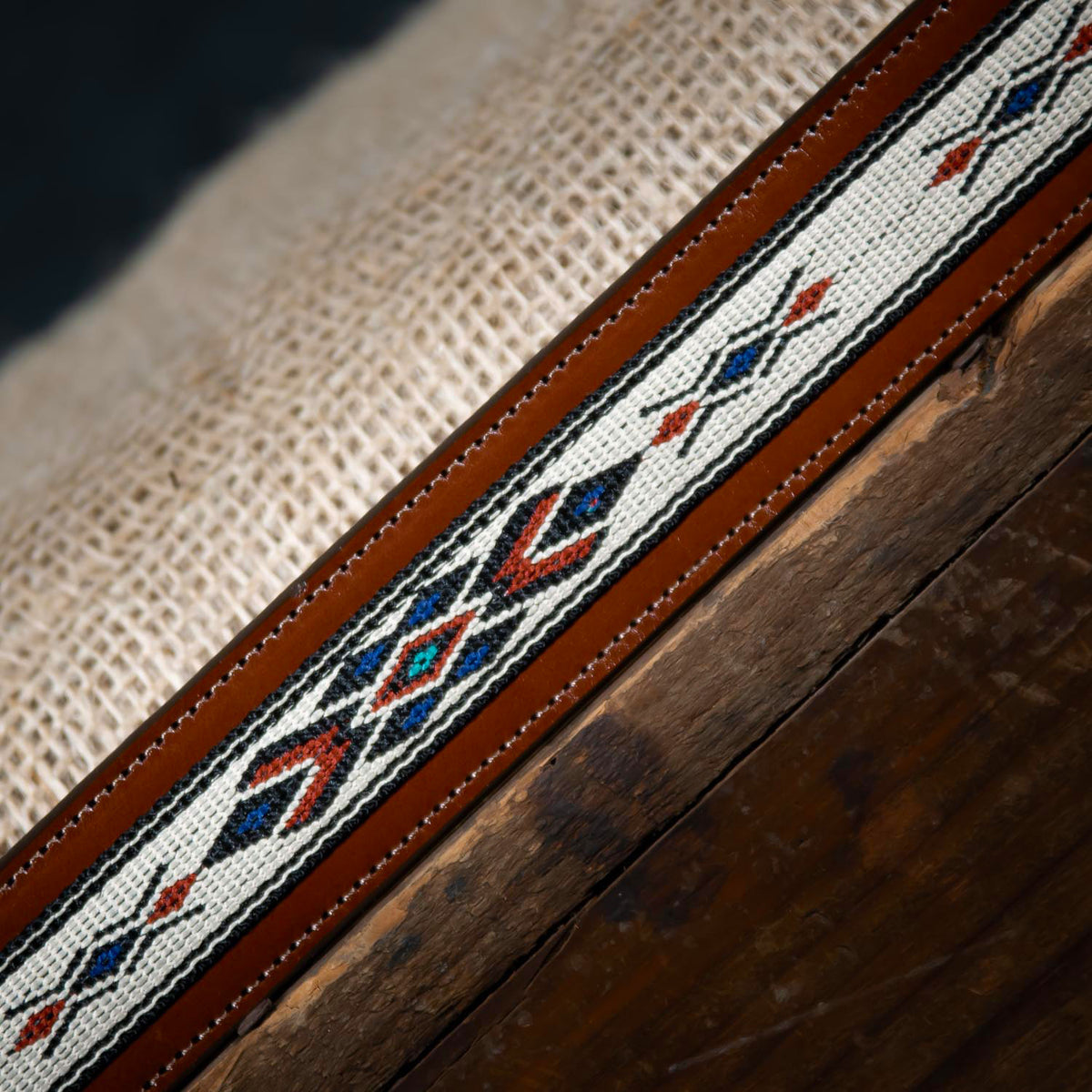 Close-up of The Seneca - 1.5&quot; by Hanks Belts, a decorative woven belt featuring intricate geometric patterns in red, blue, and black thread on a white background. The belt is finished with premium English bridle vegetable-tanned leather edges and is displayed on a textured burlap and wooden surface.