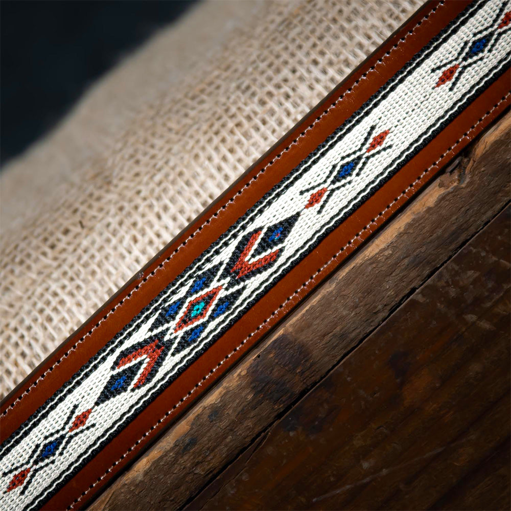 A close-up showcases The Seneca - 1.5&quot; by Hanks Belts, a brown leather belt made from premium English Bridle Leather with diamond-shaped, multicolored geometric embroidery. It exudes western spirit, resting on textured burlap on a dark wooden surface.