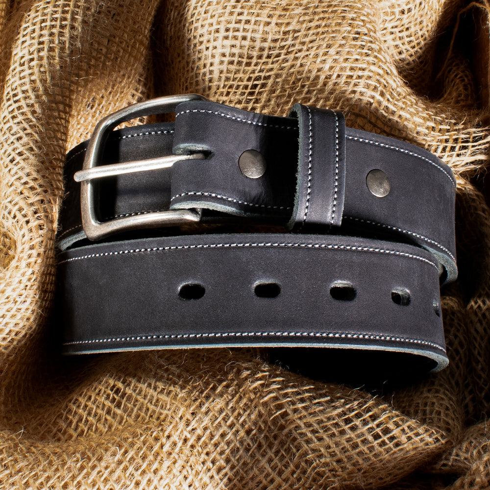 Elegant close-up of Hanks Belts&#39; USA-made &quot;The Suede Belt - 1.5&quot;&quot; in black, featuring a silver buckle, white stitching, and multiple holes. The belt is gracefully coiled on burlap fabric with sunlight casting shadows across the scene.