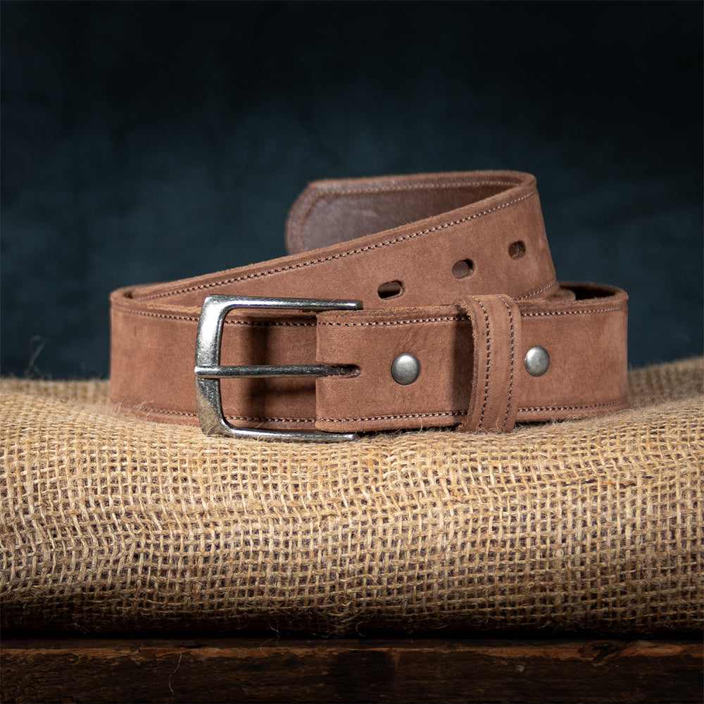 The Suede Belt - 1.5&quot; from Hanks Belts, made in the USA, features brown leather and a silver buckle, coiled on burlap against a dark backdrop to highlight its exquisite craftsmanship.