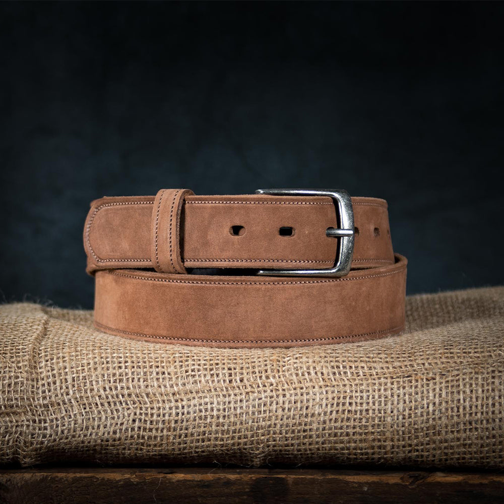 The Suede Belt - 1.5&quot; by Hanks Belts, made in the USA from fine nubuck leather, is neatly coiled on burlap. It features a silver buckle and adjustable sizing with multiple holes. A dark background highlights the rich textures of both belt and cloth.