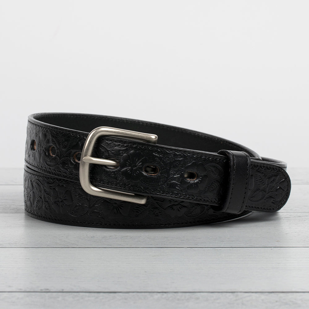 Hanks Belts&#39; Floral Belt - 1.25&quot; features black full-grain leather with an embossed floral design and a chic silver buckle, elegantly coiled on a light wooden surface.