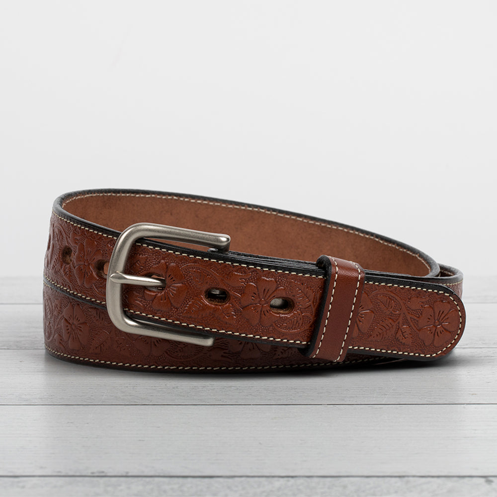 The Hanks Belts Floral Belt - 1.25&quot;, made from full-grain leather, showcases an elegant floral design with a silver buckle and white stitching, all displayed on a gray wooden surface.