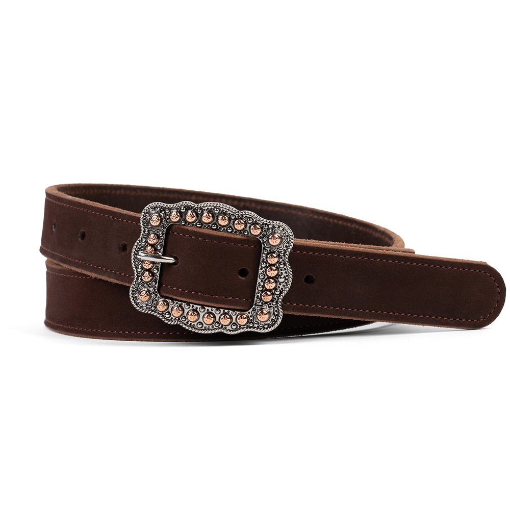 A person in white pants and a white top wears the Hanks Belts Lennon Belt - 1", featuring an ornate rose gold buckle, holding a brown wide-brimmed hat. The focus is on the soft suede leather belt and its standout buckle.