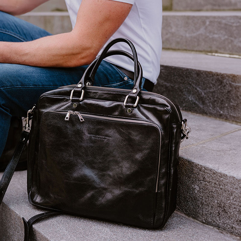 A person seated on stone steps, wearing a white t-shirt and blue jeans. Next to them on the steps is a large, black leather briefcase with two top handles, a front zippered pocket, and a shoulder strap draped to the side.