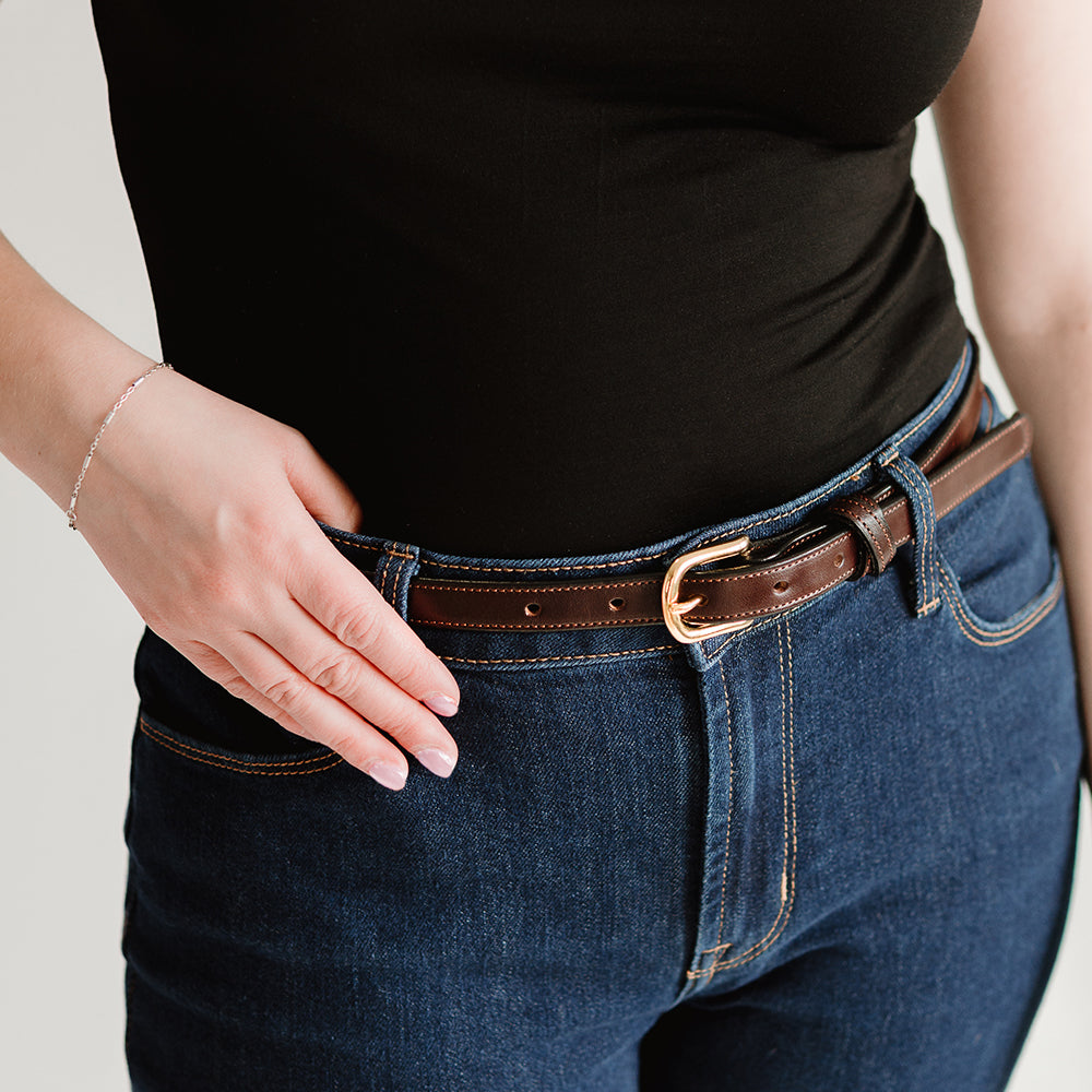 A person in a black shirt and blue jeans wears the Dixie Belt - .75&quot; in Havana by Hanks Belts, made with premium English bridle leather and adorned with a gold buckle. They rest their hand on this versatile women&#39;s accessory.