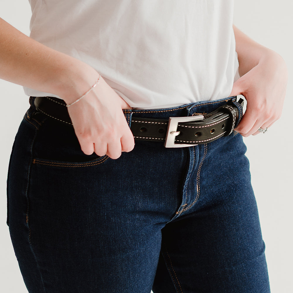 A person wearing a white t-shirt, dark blue jeans, and a USA-made 1.25&quot; Bonnie Belt by Hanks Belts, crafted from full-grain vegetable-tanned leather with a silver buckle. They are slightly adjusting the belt with both hands.