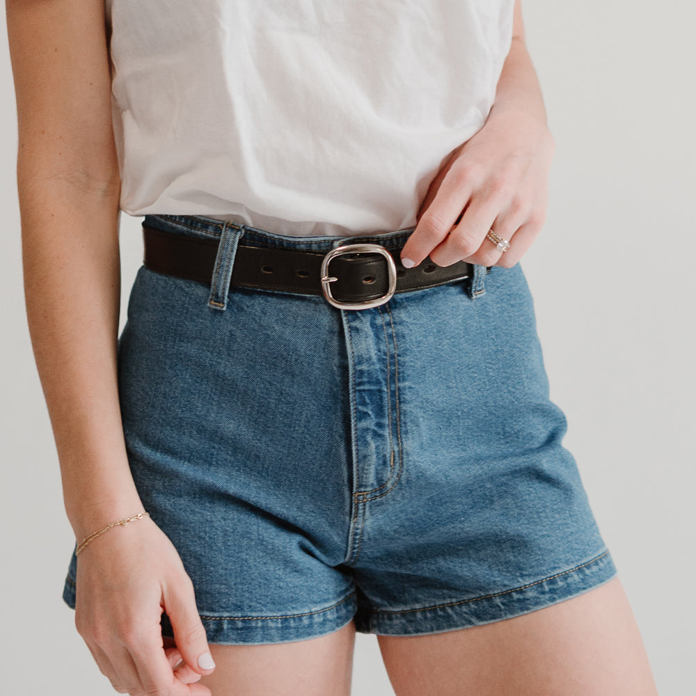 Against a plain background, a person wears high-waisted denim shorts paired with the Eloise Belt - 1.25&quot; by Hanks Belts, made in the USA from vegetable-tanned leather. The stylish black belt enhances their outfit.