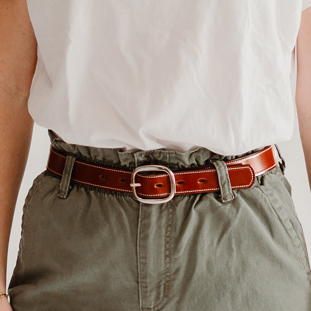 A person wears a white shirt tucked into high-waisted olive green pants, secured with the Hanks Belts Eloise Belt - 1.25&quot; in Oak, crafted from USA-made vegetable-tanned leather with a silver buckle, focusing on the waist to highlight the belt and casual outfit style.