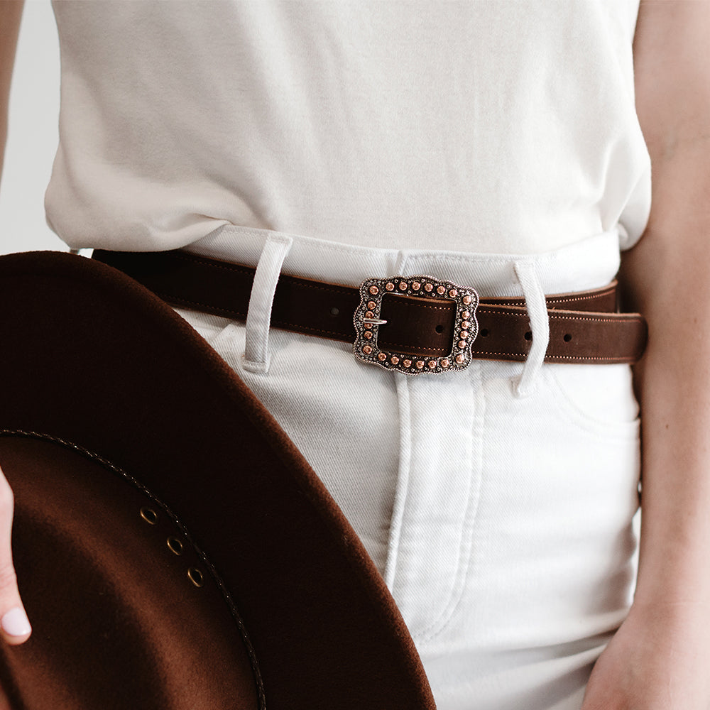 A person in white pants and a white top wears the Hanks Belts Lennon Belt - 1", featuring an ornate rose gold buckle, holding a brown wide-brimmed hat. The focus is on the soft suede leather belt and its standout buckle.