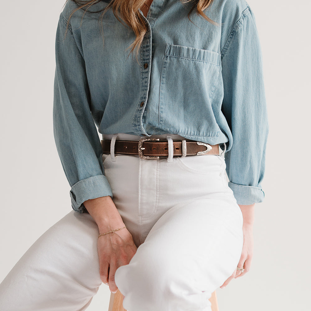 A person in a denim shirt sits on a stool, wearing high-waisted white pants and showcasing Hanks Belts&#39; Willow Belt - 1.25&quot; with its floral buckle set. Their hands rest on their lap against a plain, light-colored background.