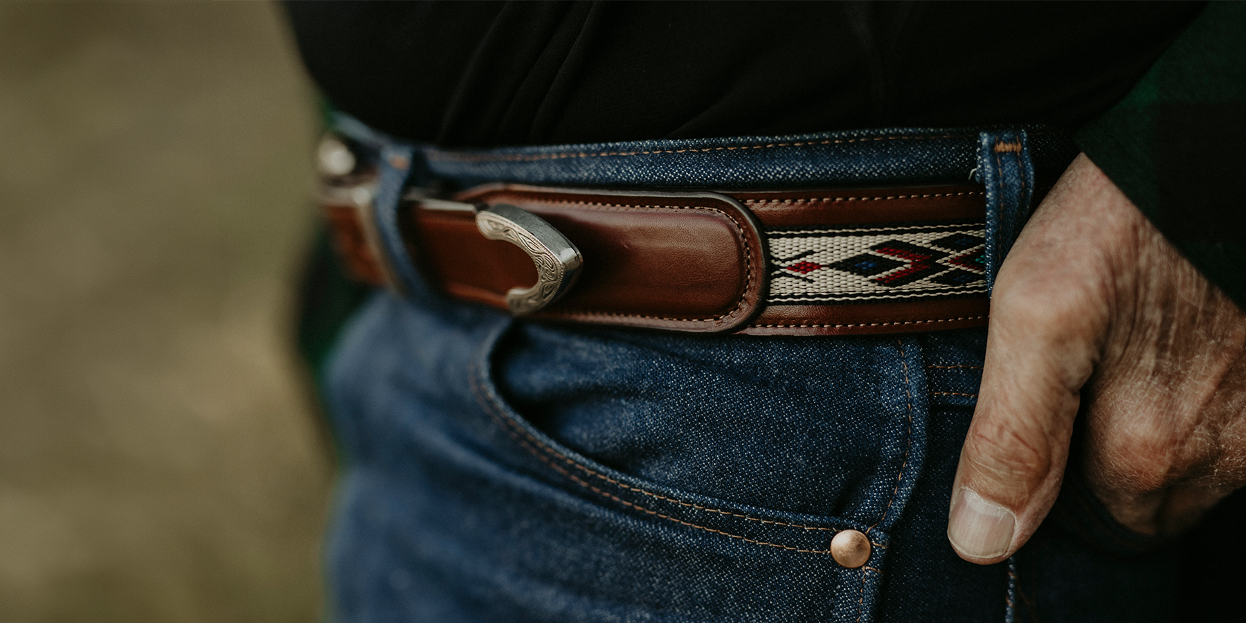 A close-up of a person wearing blue jeans with a hand in the pocket. The focus is on the brown leather belt with a silver buckle and a decorative woven pattern in red, white, and black. The person's thumb is tucked into the pocket.