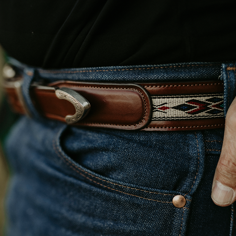 A close-up image of a person wearing blue jeans with a brown leather belt featuring a decorative buckle and an embroidered pattern. The person's hand is partially visible, resting on the right side of their jeans' pocket.