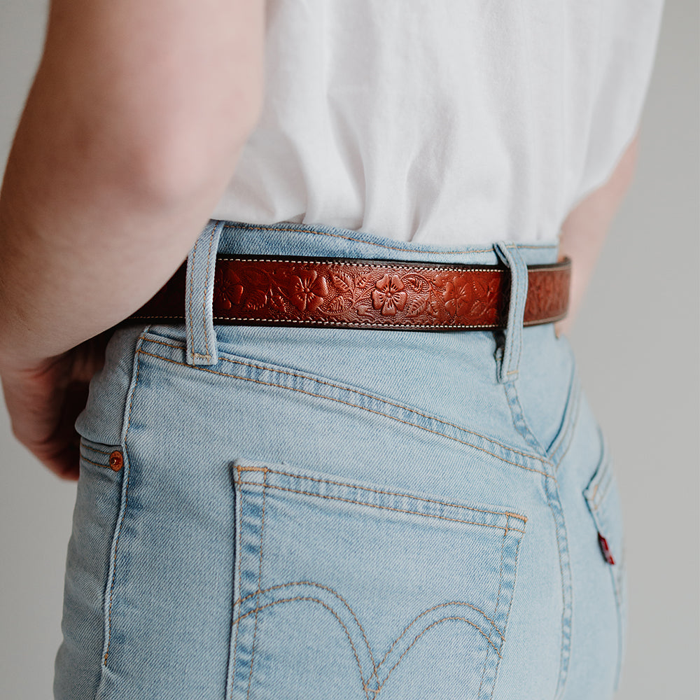 A person is seen wearing light blue jeans and a white t-shirt, with the focus on the intricate details of their outfit from the back, featuring a Floral Belt - 1.25&quot; in Oak by Hanks Belts, crafted from USA Made full-grain leather.