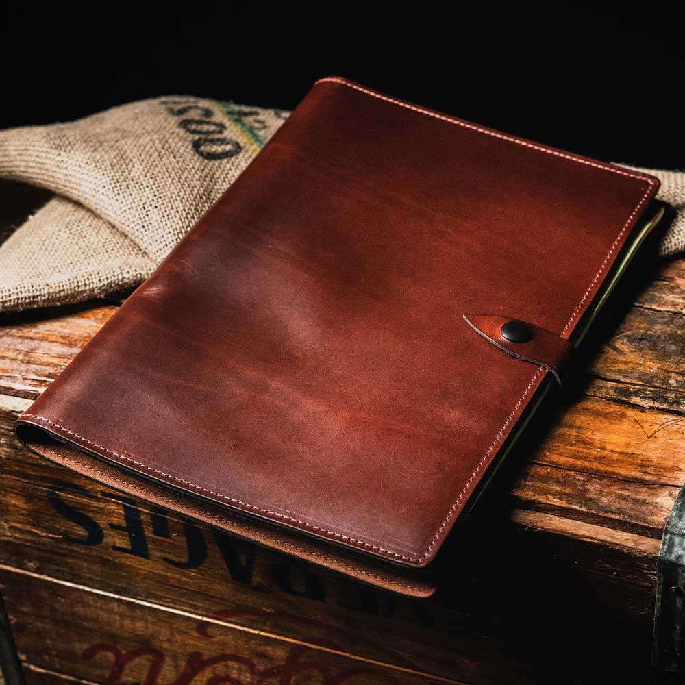 The Luxury Folio by Hanks Belts, a closed brown full-grain leather portfolio with a buttoned strap closure, is shown against a plain white background. Made in the USA, this high-end folio boasts a smooth surface and visible stitching along the edges.